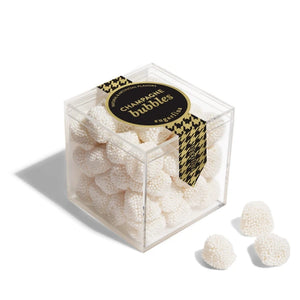 A clear acrylic cube container filled with small white champagne bubbles candy, with a black and gold houndstooth ribbon and a round black label reading 'Champagne bubbles' on top. Three individual bubbles are shown outside the container on a white background.