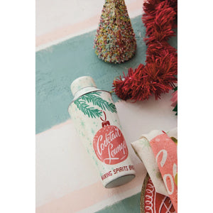 A cylindrical metal shaker, Cocktail Lounge Martini Spirits Shaker with a festive Christmas ornament label, styled on a pastel striped background with red tinsel and a miniature tree.