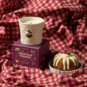 A festive Christmas Pudding scented soy wax candle by Passmanes, shown with its purple packaging box and a decorative ceramic holder. The scene is styled with a red and cream gingham check blanket, featuring a miniature pudding decoration in a white bowl.