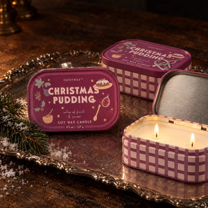 Three small rectangular tin candles labeled 'Fragrance of Christmas Pudding' are arranged on an ornate silver tray. One tin is open and lit, showing two burning wicks and cream-colored wax, while the other two tins are closed.