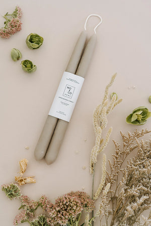 Two clay colored beeswax taper candles with a white label surrounded by dried flowers on a beige background.