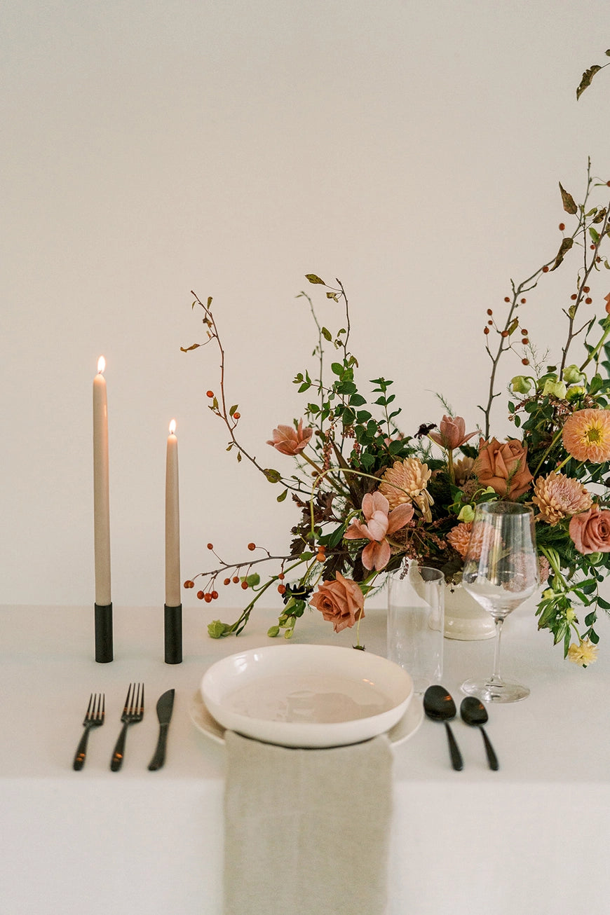 Elegant table setting with floral arrangement, clay colored beeswax taper candles, and cutlery on a white surface.