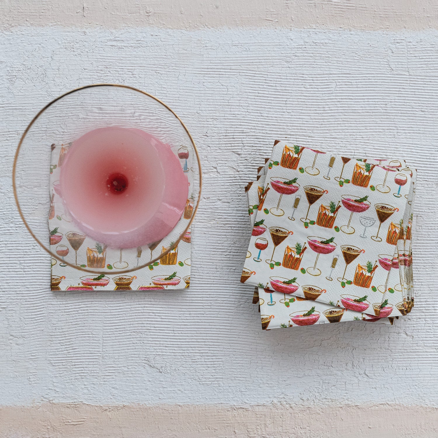A set of cocktail-themed paper napkins with a white background and colorful printed drinks, shown with one napkin folded under a clear glass coaster and a stack of folded napkins on a textured white surface.