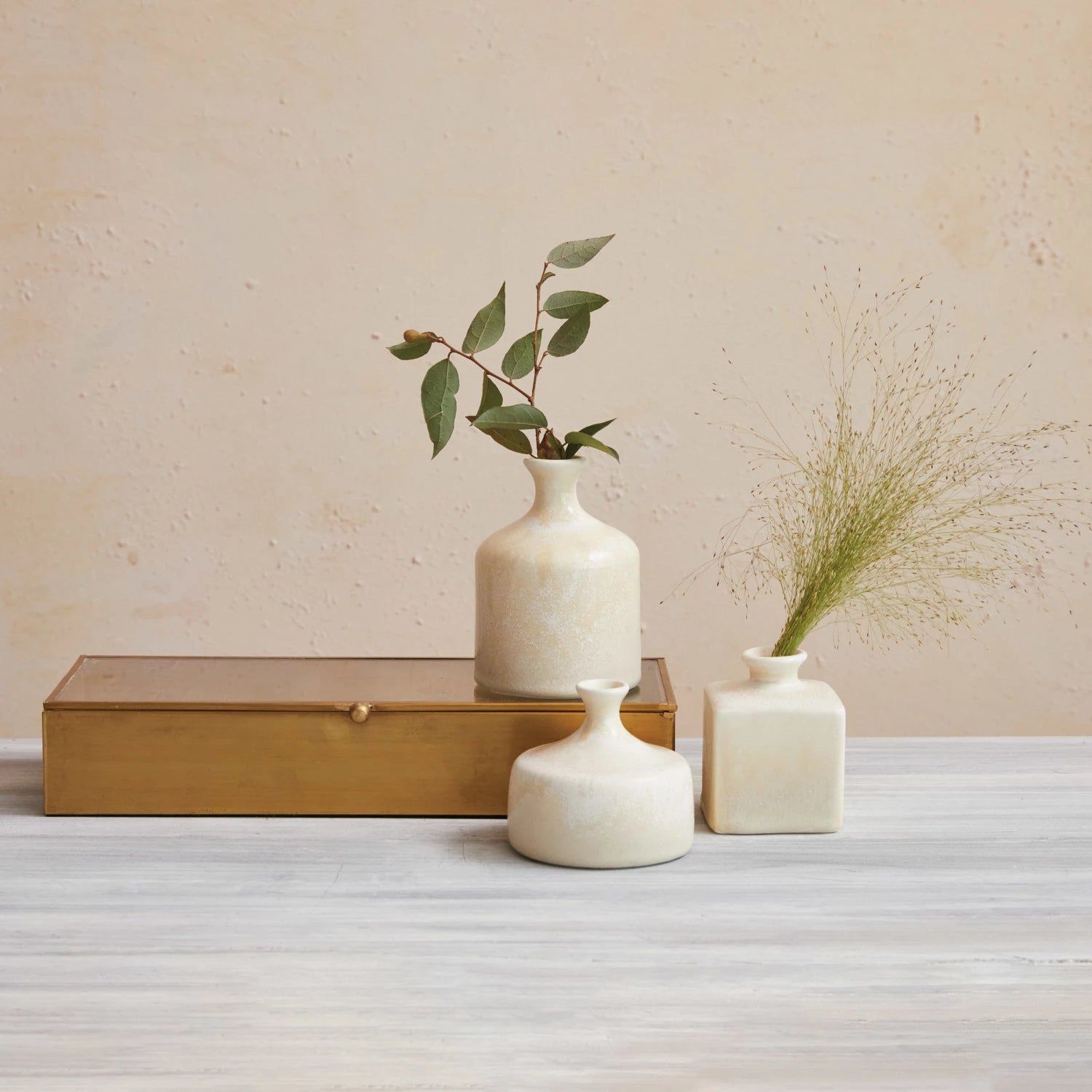 Three coordinating stoneware vases filled with plant stems on a gold metal box against a beige wall.