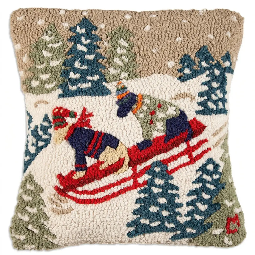 A square hook-and-loop decorative pillow featuring a winter scene with two dogs in winter hats and sweaters, sledding on snow, surrounded by pine trees, on a beige background.