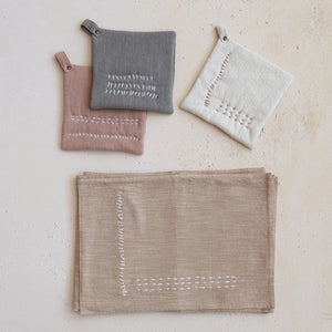Embroidered cotton slub pot holders in grey, linen, and washed terracotta. Pictured next to embroidered beige kitchen towels