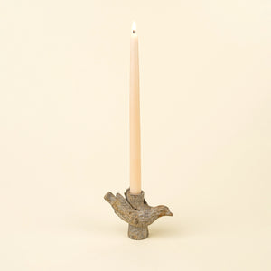 A lit taper candle in a textured bird-shaped holder, set against a solid cream background.