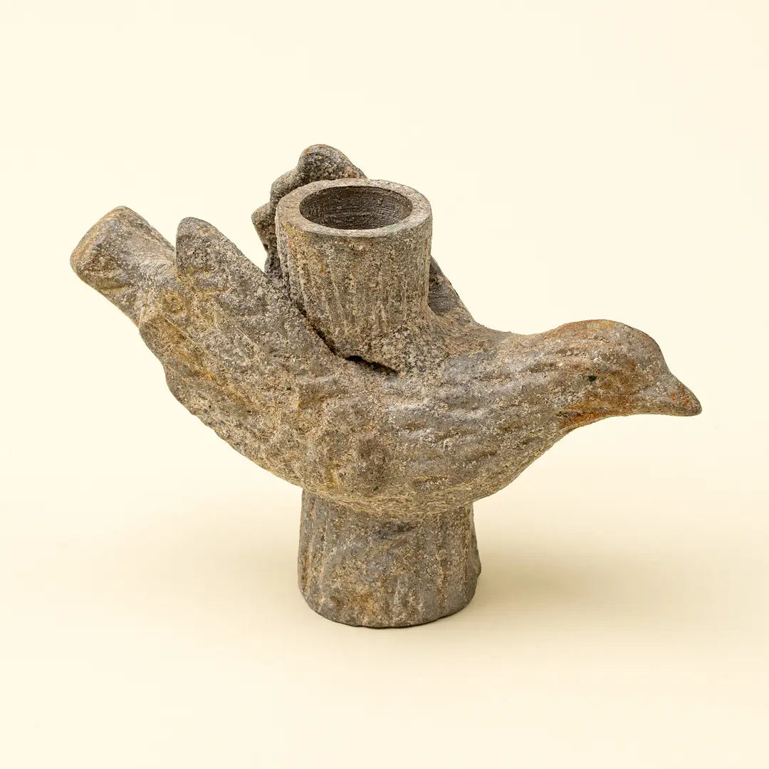 A rustic stone bird candle holder with a textured gray-brown finish, featuring a bird body with wings folded back to form the candle holder's cup, set against a solid cream background.