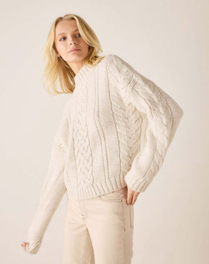 A blonde woman wearing an oversized cream cable-knit sweater and light beige pants, photographed against a plain light beige studio background.