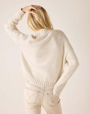 A woman shown from behind wearing a cream-colored, chunky knit turtleneck sweater and light beige jeans against a solid light beige background.