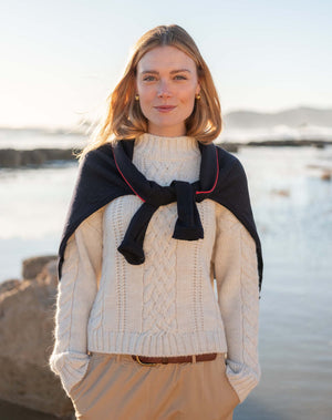 A woman standing outdoors by the water wearing a cream cable-knit sweater, beige pants, and a black shawl draped over her shoulders with thin red piping.