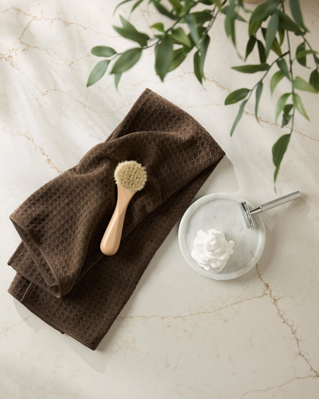 Brown towel with a brush and soap dish on a marble surface with green leaves.