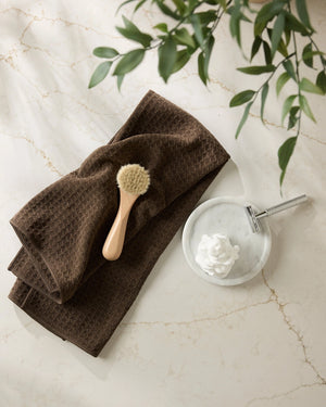 Brown towel with a brush and soap dish on a marble surface with green leaves.
