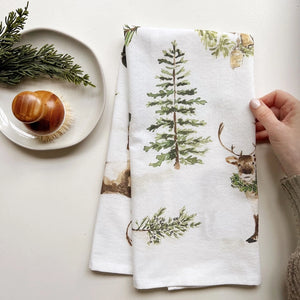 A white cotton tea towel with Christmas greenery illustrations, including trees and a reindeer, displayed on a white surface with a piece of greenery in the background and a dish brush in the foreground. Emily Lex Studio. 