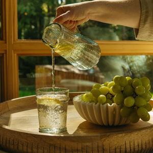 Person pouring water from a glass pitcher into a glass on a wooden table with grapes in the white fluted catchall bowl.