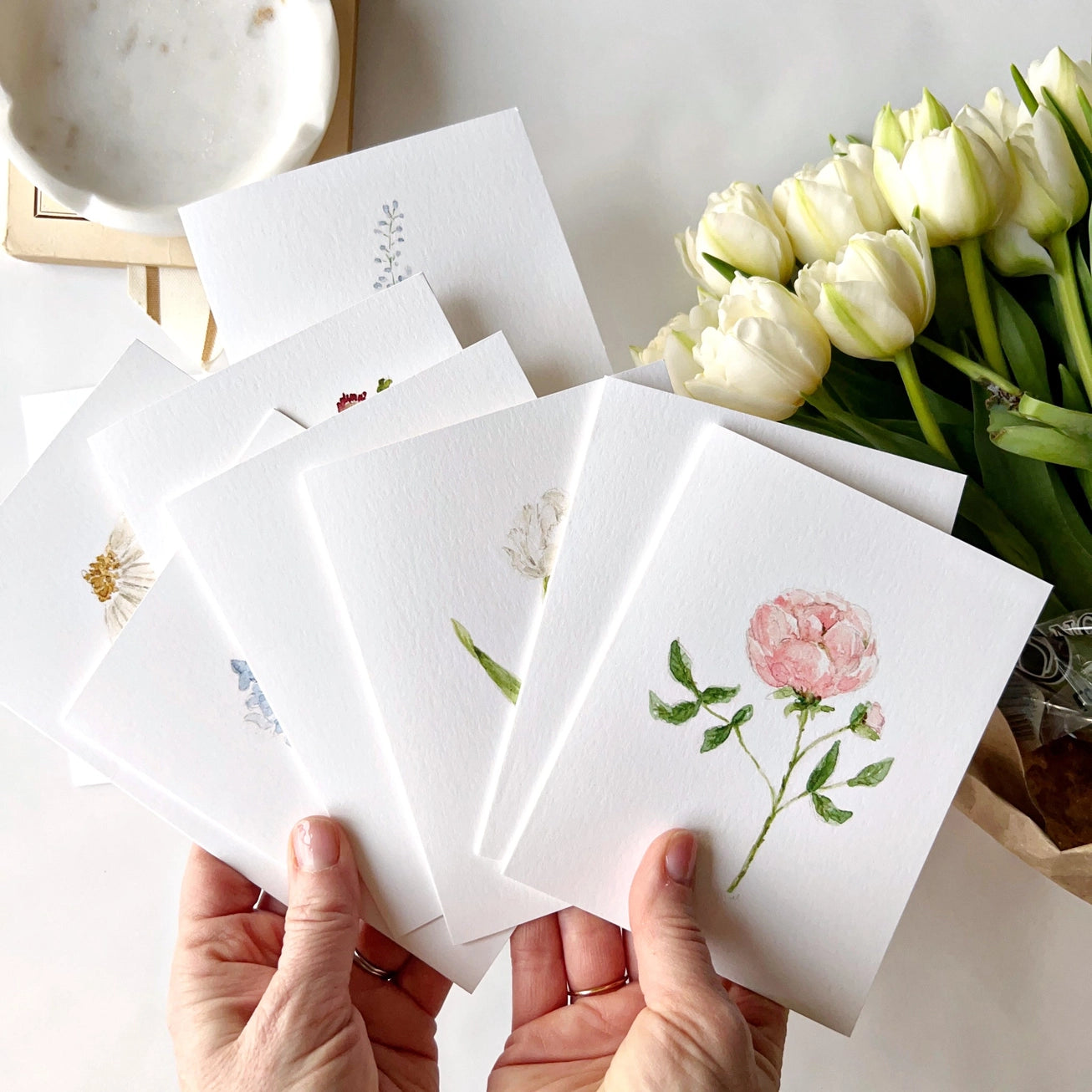 Set of floral cards held by hands with a bouquet of white tulips in the background. Emily Lex notecard set. 