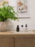 Three L'Avant mini bottles next to each other on a lucite tray and near a large green plant., dish soap, hand soap, and multicleaner spray.