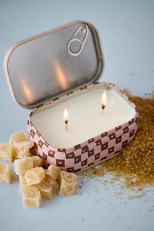 A two-wick candle in an open tin container with a pink and brown checkered pattern, styled with sugar cubes and loose sugar on a light blue background.