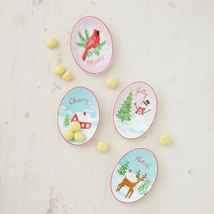 Four oval Christmas-themed candy dishes arranged on a light off-white wooden surface with small yellow candies scattered around them. The dishes feature festive illustrations: a red cardinal on pine branches with the word Merry, a snowy house with the word Cheery, a snowman beside a Christmas tree with the word Jolly, and a brown deer with the word Noel.