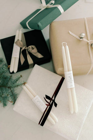 Ivory taper candles with labels on surface with wrapped gifts in the background. Beeswax candles. 