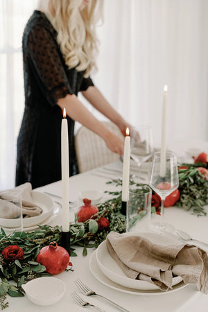Table setting with beeswax taper candles, pomegranates, and greenery on a white tablecloth. 