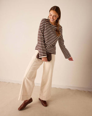 A woman modeling a brown and cream striped long-sleeve top with wide-leg cream pants and brown loafers, standing against a plain beige studio wall.