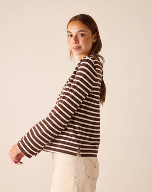 A woman wearing a brown and cream striped long-sleeve knit top, shown from a side-back angle against a plain beige studio background.