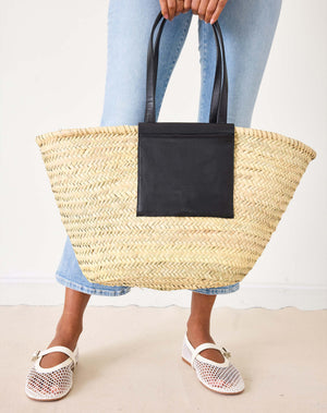 Person holding a natural straw tote bag with a black leather pocket patch against a white background. 