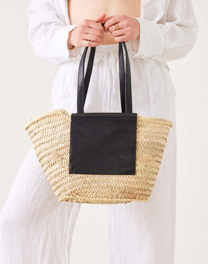 Person holding a straw tote bag with black leather top handle against a white background. 