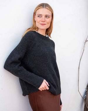 A woman with blonde hair wearing a dark charcoal gray ribbed knit sweater and brown skirt, leaning against a white wall.