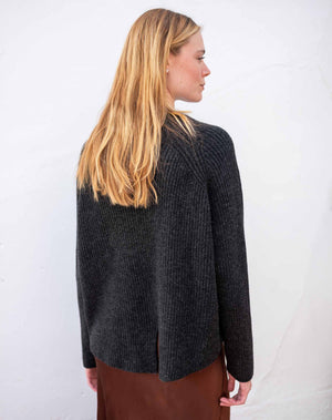 A woman with long blonde hair is shown from the back wearing a dark charcoal gray ribbed knit sweater and a brown skirt, standing against a white wall.