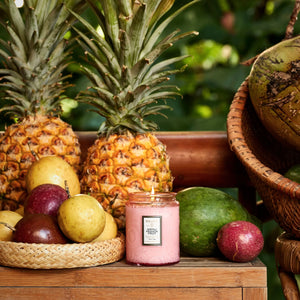 Pink candle with a label surrounded by pineapples, passion fruits, and other fruits on a wooden surface.
