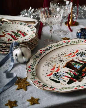 A festive holiday table setting featuring a stack of white ceramic plates with colorful illustrations of the nativity, a silver ornament, clear glass goblets, and gold star decorations on a gray tablecloth.