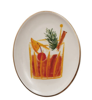 An oval white ceramic plate with a gold rim, featuring a hand-painted illustration of a winter cocktail with an orange slice, cinnamon stick, pine sprig, and a red cherry garnish.