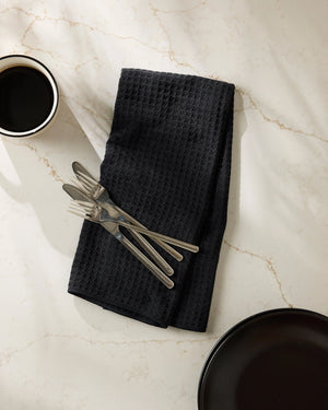 Black waffle-textured tea towel with silverware on a marble surface with a black plate and cup.