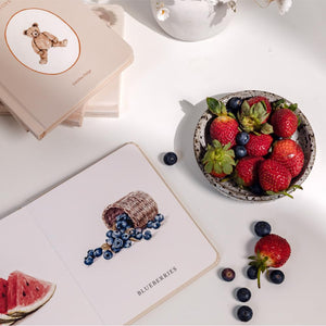 Fruits and berries on a white surface with a notebook featuring illustrations of blueberries and strawberries.