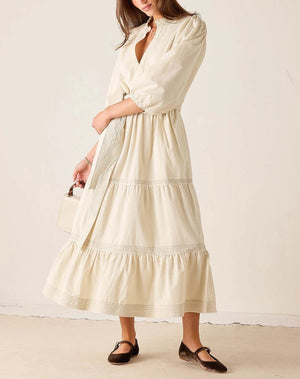 A woman wearing a cream-colored, tiered midi dress with puffed sleeves and a wrap-style front, paired with dark brown flat shoes and holding a small white handbag, set against a plain beige wall and floor.