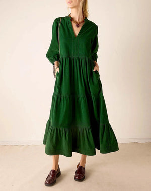 A woman wearing a dark green tiered midi dress with long sleeves, a V-neckline, and side pockets, paired with dark brown leather loafers, standing against a plain beige wall.