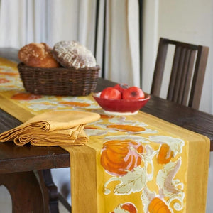Mustard yellow, orange, and gold table runner with pumpkin and autumn florals on a wooden table with bread and apples. Couleur Nature. 