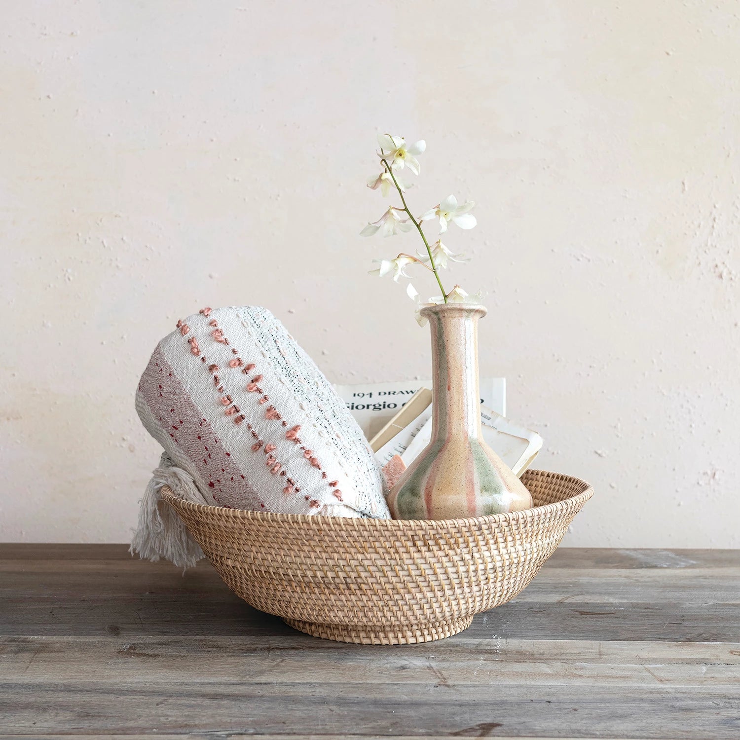 Hand woven natural rattan footed bowl with blanket and vase displayed inside. 