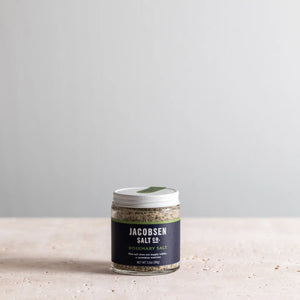 Jar of Jacobsen Salt Co. rosemary sea salt on a light surface with a gray background