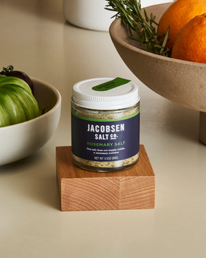 Jar of Jacobsen Salt Co. rosemary salt on a wooden block with bowls of oranges and rosemary in the background.