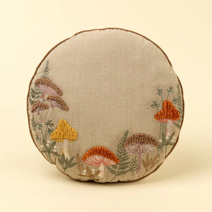 Round embroidered pillow with mushroom design on a beige background. 