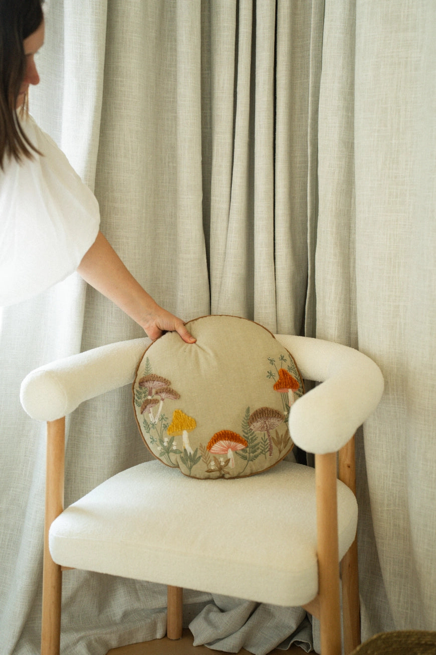 Person holding a round embroidered mushroom pillow on a chair with curtains in the background.