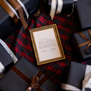 A small ornate gold picture frame holding a cream-colored card with printed text, placed on top of a red and black plaid fabric and surrounded by dark wrapped gift boxes with ribbons.