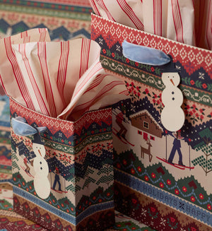 Two Christmas-themed gift bags filled with red and white striped tissue paper, set against a patterned holiday surface. The bags feature a folk-art winter scene with skiers, a cabin, a snowman, and festive borders.