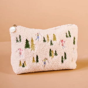 A cream-colored sherpa zippered pouch decorated with embroidered pine trees and small skiers, shown on a solid peach background.