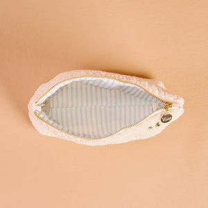 A plush, fuzzy cream-colored zippered pouch with a gold zipper and a light blue and white striped interior, shown open on a solid peach background.
