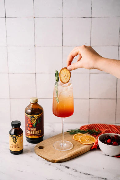 Person garnishing a spiced cranberry cocktail with a slice of fruit, with bottles and ingredients on a marble surface.