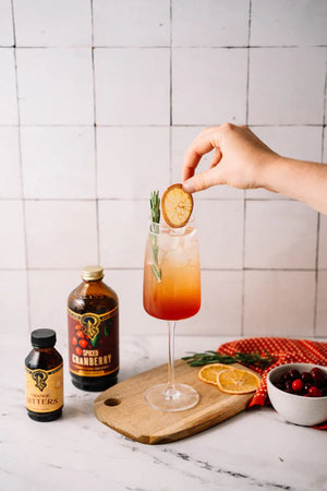 Person garnishing a spiced cranberry cocktail with a slice of fruit, with bottles and ingredients on a marble surface.